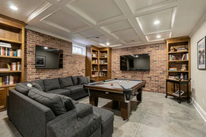 Cave transformée en salle de loisirs avec canapé en tissu gris, table de billard et mur en briques apparentes